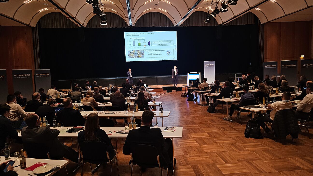 Markus Rössinger und Tobias Wallrapp auf der Bühne. Teilnehmer im Konferenzraum beim Deutschen Fachkongress Fabrikplanung 2025 im Vordergrund. 