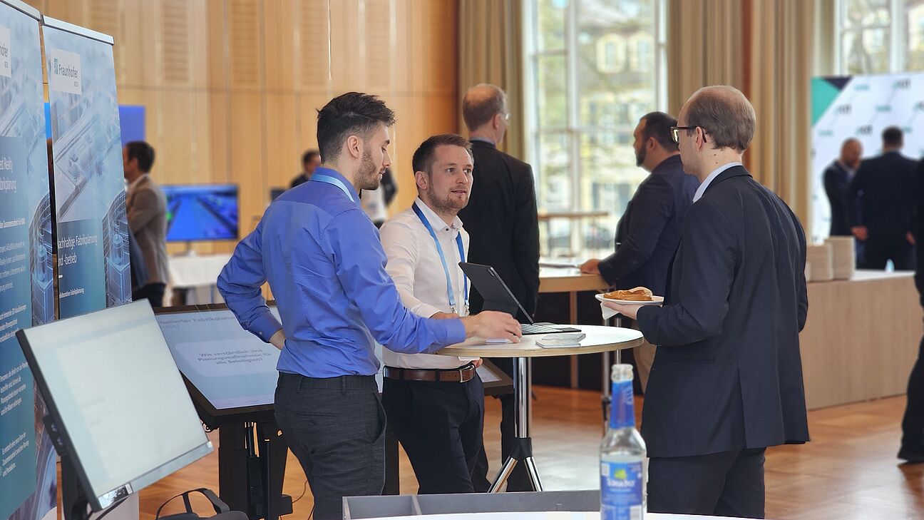 Drei Teilnehmer im Vordergrund am Stand von Fraunhofer IGCV am Netzwerken im Ausstellerbereich des Deutschen Fachkongress Fabrikplanung 2025