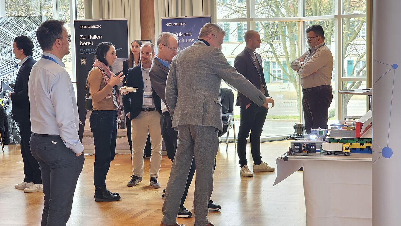 Teilnehmer im Ausstellerbereich des Fachkongress Fabrikplanung 2025 am Netzwerken. Im Hintergrund sieht man den Stand von GOLDBECK
