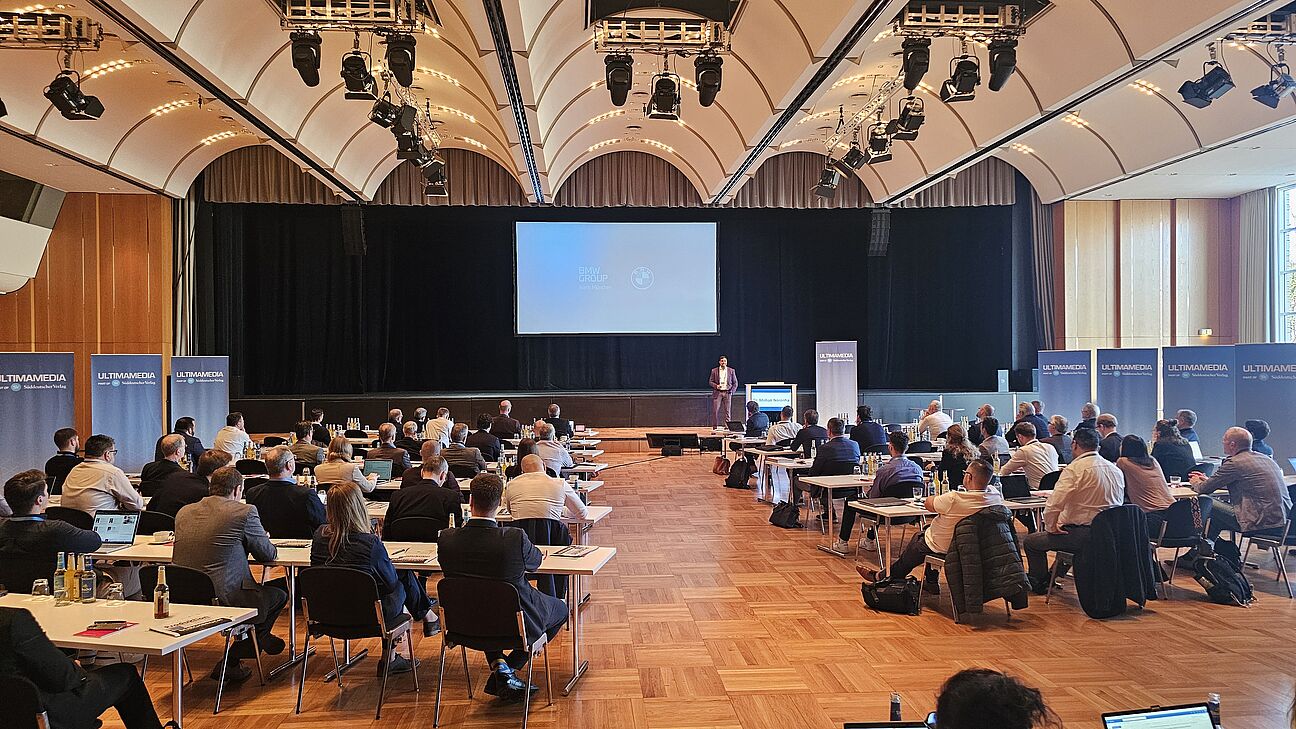 Dr. Mohan Noronha auf der Bühne. Man sieht die Teilnehmer im Vordergrund im Konferenzraum beim Fachkongress Fabrikplanung 2025