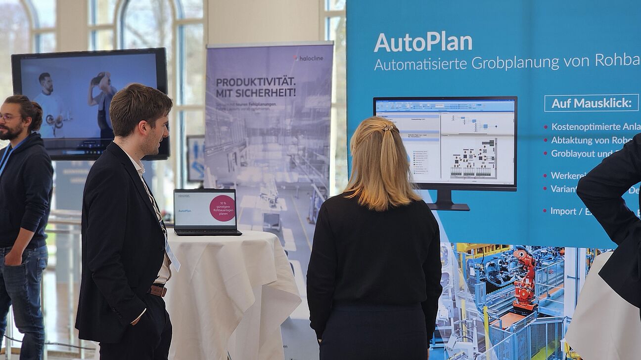 Zwei Teilnehmer am Netzwerken auf dem Deutschen Fachkongress Fabrikplanung 2025. Man sieht die Stände von halocline und adalogic.